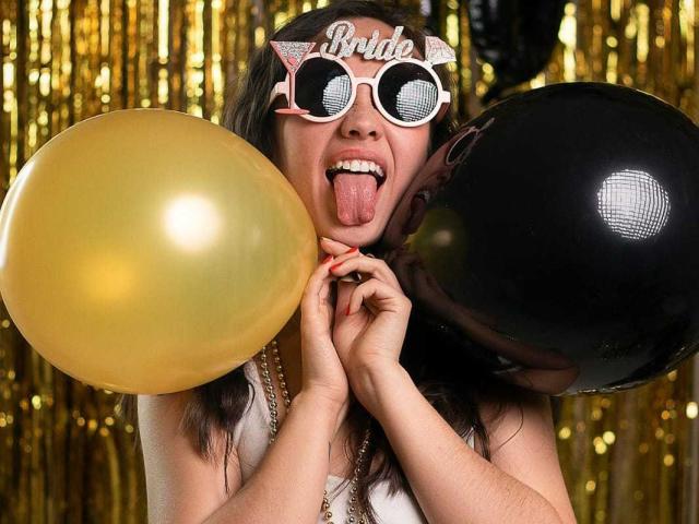Lila69Hot with pink glitter "Brute" glasses, tongue out, holding gold and black balloons, wearing white top, against golden streamers. Her tits are partially visible.