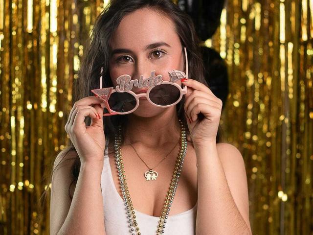 Lila69Hot with pink "Bitch" sunglasses, holding them in front of her face, wearing a white tank top, gold necklace, and a elephant pendant, against a gold curtain.