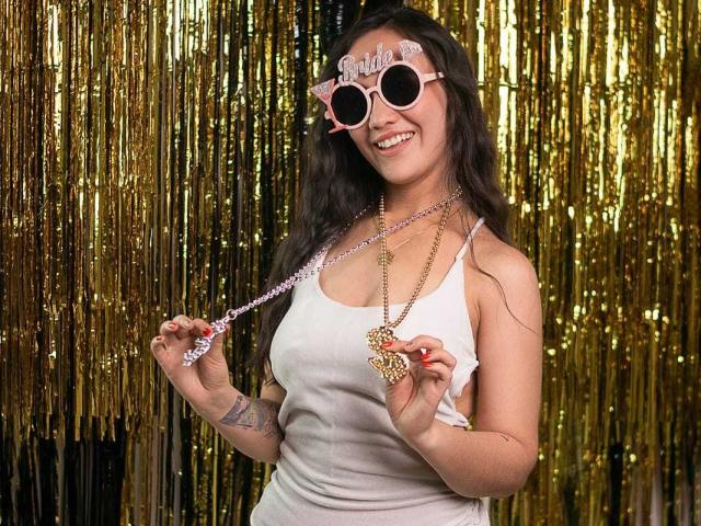 Lila69Hot with pink cat-eye glasses, white tank top, holding pink beaded necklace and gold heart, smiling, has tattoos on left arm, wearing gold chain, brown hair, gold curtain.