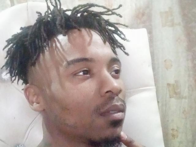 TaurusBallz heeft donkerbruine huid, korte zwarte dreadlocks, een witte verbanddoek op zijn voorhoofd en een dunne zwarte baard. Hij zit in een witte stoel en kijkt weg..