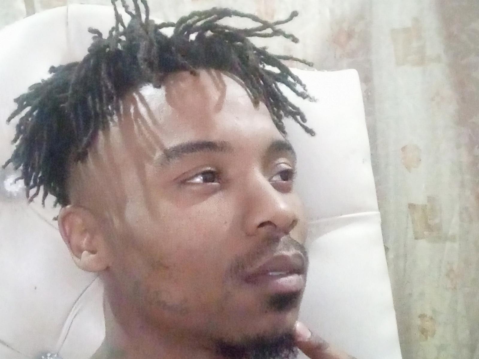 TaurusBallz heeft donkerbruine huid, korte zwarte dreadlocks, een witte verbanddoek op zijn voorhoofd en een dunne zwarte baard. Hij zit in een witte stoel en kijkt weg..
