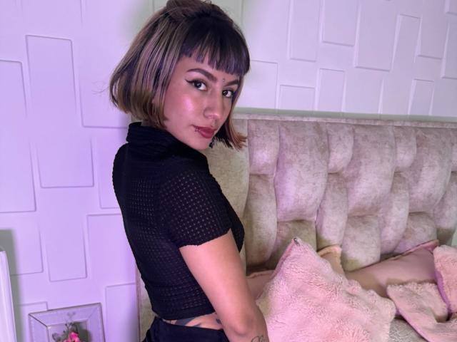 AmberTowsend with short brown bob, black mesh top, side glance, pinkish skin, subtle makeup, sitting on beige tufted couch, pink fluffy pillow, white wall, slightly smiling.