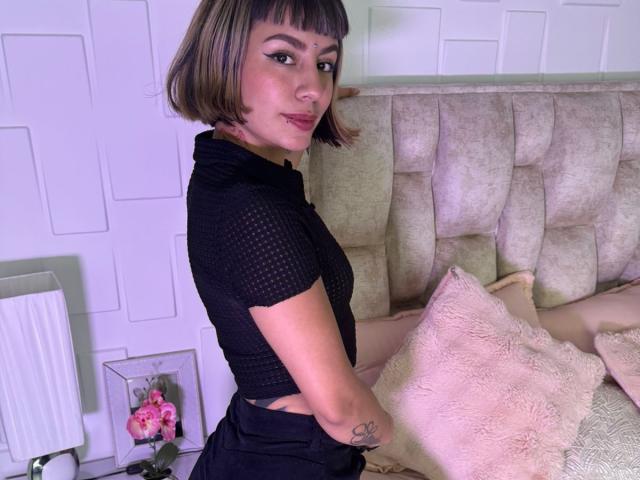 AmberTowsend with short brown bob, black mesh top, black skirt, tattoo on forearm, pink pillow, beige headboard, white wall, standing sideways, looking over shoulder, pink lipstick.