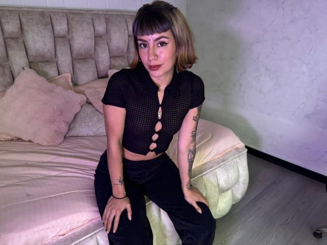 AmberTowsend with short brown hair, wearing a black mesh top with button holes, black pants, and sitting on a bed with pink pillows, showing tattoos on her arms.