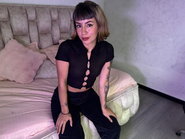 AmberTowsed, with short blonde hair, sits on a bed in black mesh top with cut-out, showing small tits, black pants, tattoos on arms, and pink pillows in background.