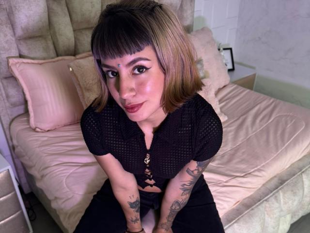 AmberTowsend, with short brown hair and tattoos, wears a black lace-up top, sitting on a bed with pink sheets, looking up with a slight smile, showing her tits.