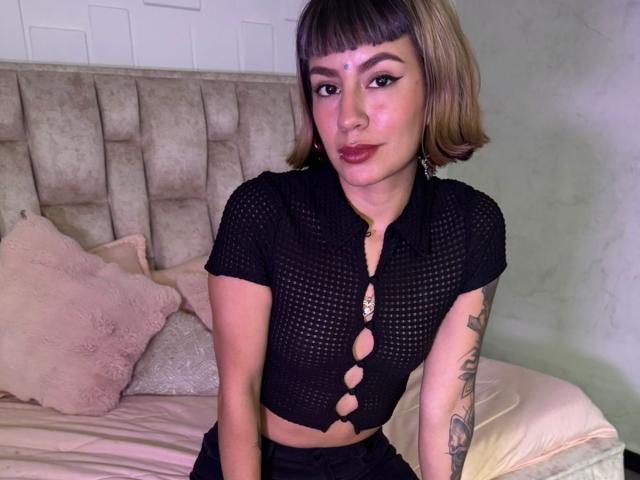 AmberTowsend with short brown bob, black mesh crop top showing tits, button cutouts, tattooed arm, pink pillows, beige headboard, and red lipstick, sitting on bed.