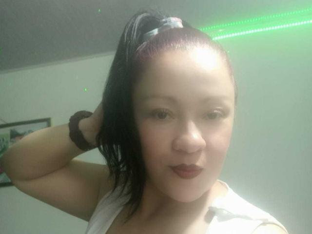  CharloteEzra with dark hair in a ponytail, holding her hair up, wearing a white top, and a dark scrunchie, has red lipstick and a slight smile, looking at camera.