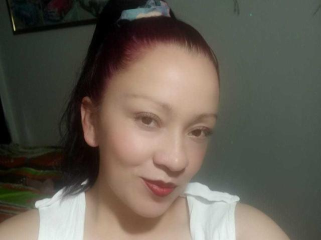 CharloteEzra has light skin, red lipstick, and dark red hair in a ponytail with a blue scrunchie, wearing a white top, and smiling slightly with her nose slightly up.