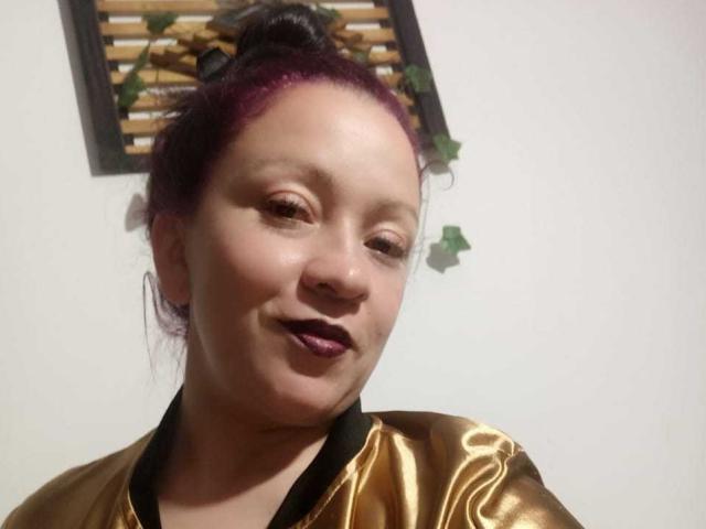 CharloteEzra has purple hair in a bun, wearing a shiny gold top, smiling with dark lipstick, and has a cute, slightly chubby face.