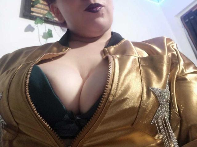 CharloteEzra with large, shiny gold jacket zipped down, exposing her big, cleavage-filled tits in a green lace bra. She's got dark purple lipstick and a slightly chubby face.