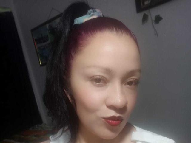  CharloteEzra with red-tinted black hair in a ponytail, light skin, subtle makeup, red lipstick, and a slight smile, wearing a white shirt, in a dimly lit room.