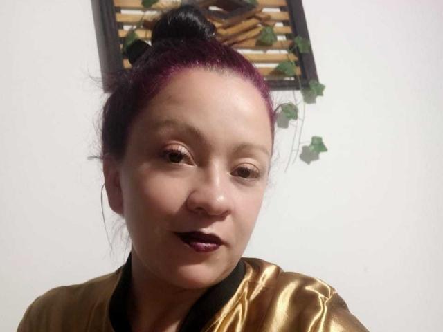 CharloteEzra with dark purple hair in a bun, wearing a shiny gold shirt, dark purple lipstick, and a slight smile, showing her face and upper chest.