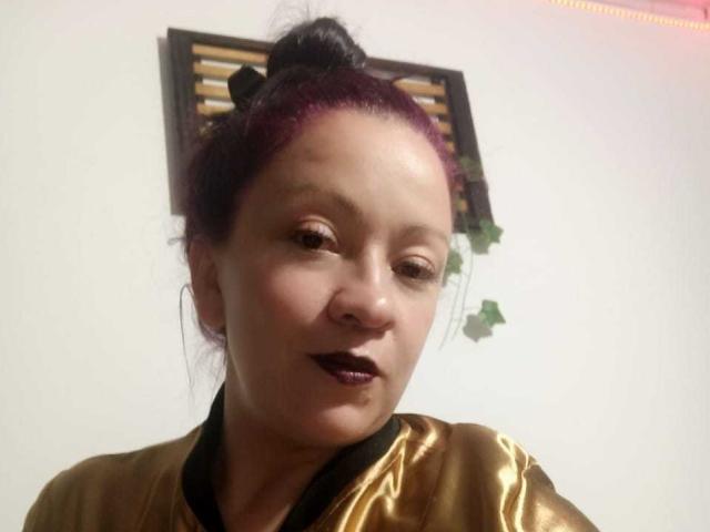 CharloteEzra with purple hair in a bun, wearing dark lipstick, gold satin robe, black collar, looking slightly to the side, medium skin tone, soft expression, slight smile.