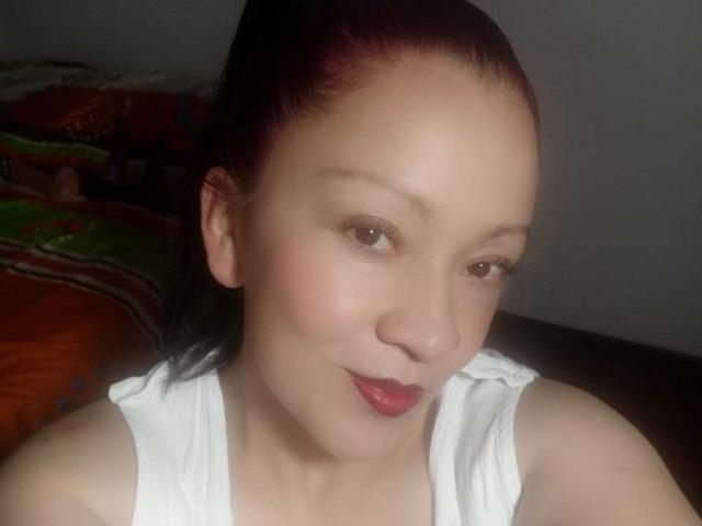  CharloteEzra with red-tinted hair, light skin, brown eyes, and red lipstick, wearing a white top, smiling softly at the camera, showing her cute, pretty face.