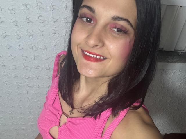 DulceSweety with pink eye makeup, red lipstick, and straight black hair, smiling at the camera, wearing a pink ribbed top with a keyhole cutout, showing her tits.