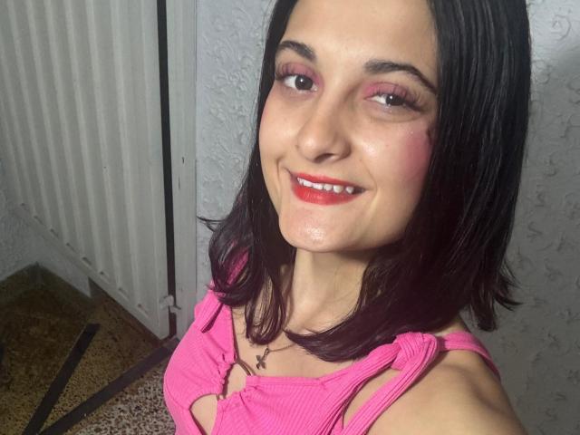 DulceSweety with dark hair, pink eyelids, red lipstick, and a pink top showing her cleavage, smiling at the camera. She's standing in a room with a textured wall.