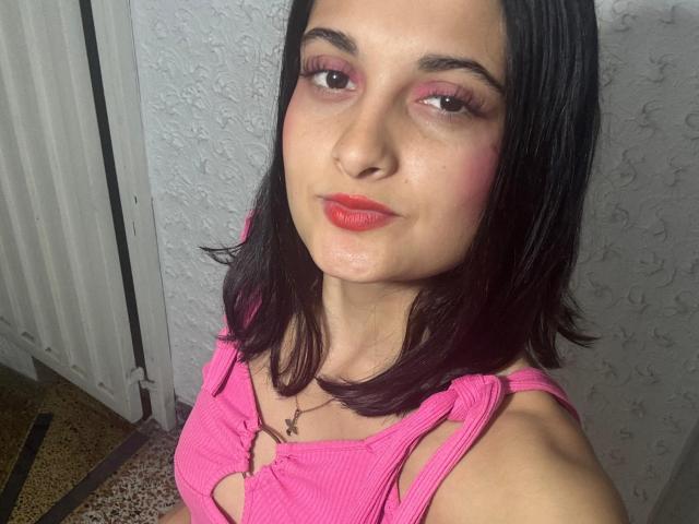 DulceSweety with pink makeup, shiny red lips, black shoulder-length hair, pink ribbed top with cutout, small breasts, and a necklace, in a selfie.
