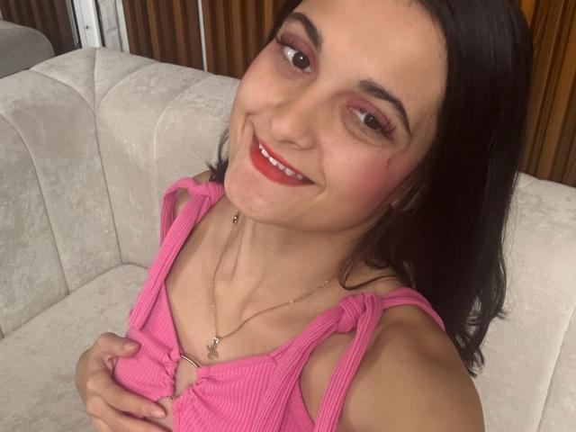 DulceSweety with pink ribbed top, holding her tits, smiling, red lipstick, brown eyes, black hair, gold necklace, cute, sitting on white couch, wood background.