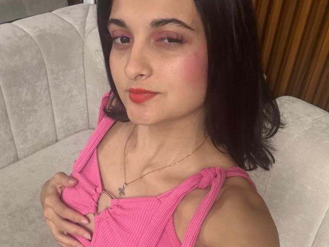 DulceSweety with pink ribbed tank top, holding her small tits, red lipstick, pink eye shadow, black hair, light skin, silver necklace, sitting on white couch, wood background.