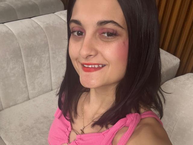 DulceSweety has pink eyeshadow, red lipstick, and a cute smile. She's wearing a pink ribbed top, showing her small tits, and has a slight blush on her cheeks.