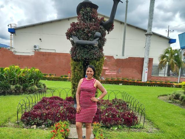 PauletteLoveX stands in front of a tall, leafy statue with a pipe on top, wearing a red and white striped dress, smiling with hands on hips, grassy background.