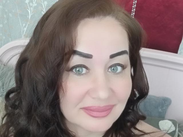 LexyRose has blue eyes, dark brown wavy hair, thick eyebrows, pink lipstick, and a slight smile. Her pale skin contrasts with her dark hair. She looks cute.