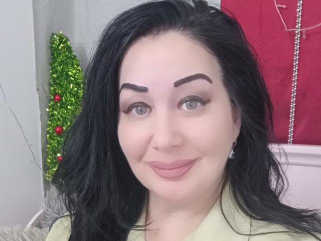 LexyRose has black hair, dark eyebrows, light skin, blue eyes, and pink lips, wearing a yellow shirt, with a small green Christmas tree and red ornaments in the background.