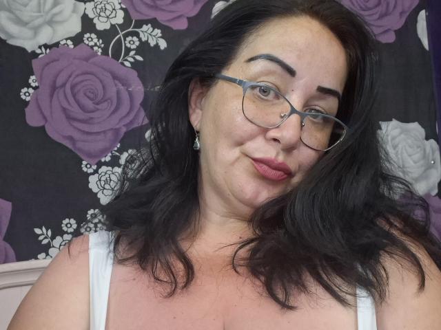 LexyRose has black hair, glasses, a nose ring, and is wearing a white top. Her pink lips are slightly puckered. Her tits are medium-sized, and she's smiling softly.
