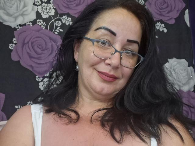 LexyRose with dark hair, glasses, nose ring, and pink lipstick, smiling at the camera, wearing a white top, showing her tits. She has a cute, sexy, and confident look.