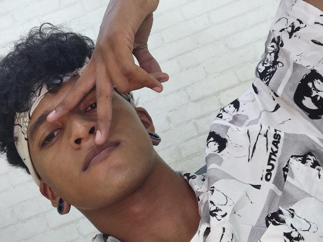 ChrisMartinez with curly black hair, wearing a white and black graphic shirt, headband, and ear gauges, covering his right eye with his finger, has a serious expression.