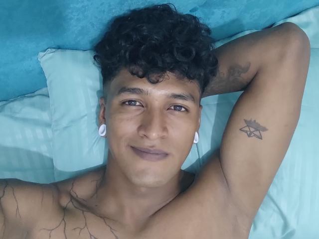 Chris Martinez, avec ses cheveux noirs bouclés, sa peau mate, ses anneaux d'oreille blancs, un tatouage sur le bras droit, allongé sur un lit, les bras derrière la tête, torse nu, souriant légèrement, regardant la caméra..