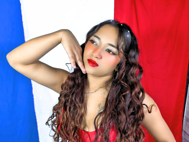 NicoleMartins with long curly brown hair, red lipstick, and shiny eyeliner, wearing a red top, hand on cheek, standing against a French flag. She has small tits and a tattoo.
