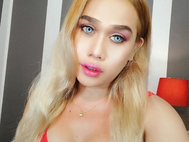RicaOnTop heeft blond haar, blauwe ogen, roze lippenstift en een gouden ketting, draagt een rode top, en er staat een rode lamp op de achtergrond. Haar borsten zijn gedeeltelijk zichtbaar..