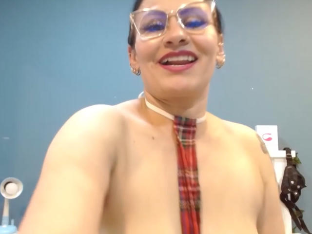 DonnaJones with glasses, red plaid tie, and no top, smiling with tongue out, showing bare tits and a choker. Her dark hair is pulled back. She's wearing red lipstick.