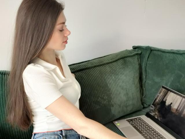EvaMcCoy with long brown hair, white ribbed shirt, and blue jeans, sitting on green couch, using silver laptop with cat on screen. Her tits are small, eyes closed.