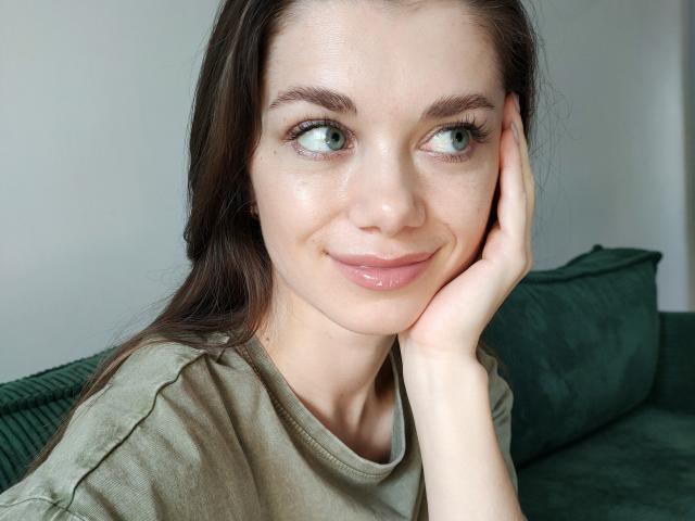 EvaMcCoy with shiny skin, green eyes, and glossy pink lips, resting her hand on her cheek, wearing a green shirt, brown hair, and a slight smile.