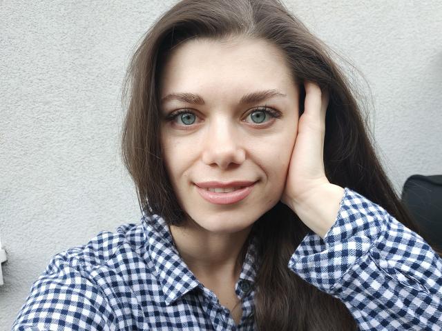 EvaMcCoy with green eyes, long brown hair, and a blue and white checkered shirt, smiling with her hand on her cheek. Her pink lips are slightly parted.