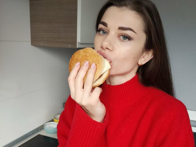 EvaMcCoy with long brown hair, red sweater, and blue eyes, eating a burger with her right hand, looking at the camera. Her lips are around the bun.