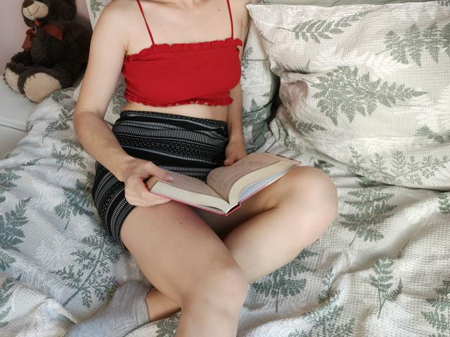 EvaMcCoy with pale skin, red crop top, and black striped shorts, sitting on a bed reading a book, her legs crossed, a stuffed bear in the background.