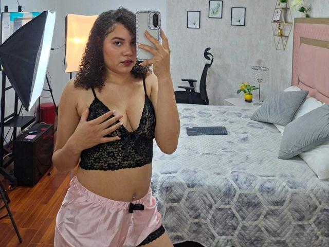 AlisonRed with curly brown hair, wearing black lace bra, pink shorts, holding phone, covering her tits with hand, standing in bedroom, wooden floor, bed with gray patterned blanket.