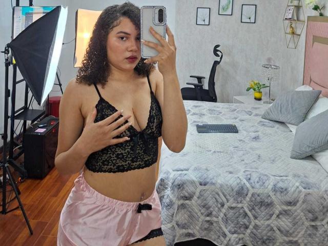 AlisonRed with curly brown hair, wearing black lace bra, pink satin shorts, holding phone, covering small tits with hand, standing in bedroom with bed and photography lights.