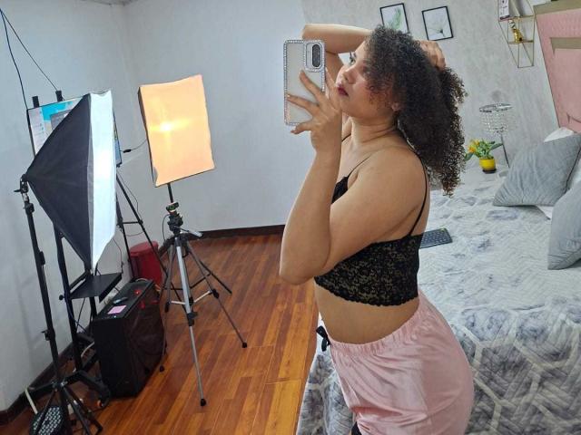 AlisonRed with curly hair, black lace bra, pink shorts, taking a mirror selfie in a bedroom, holding a phone, standing in front of two lit studio lights.