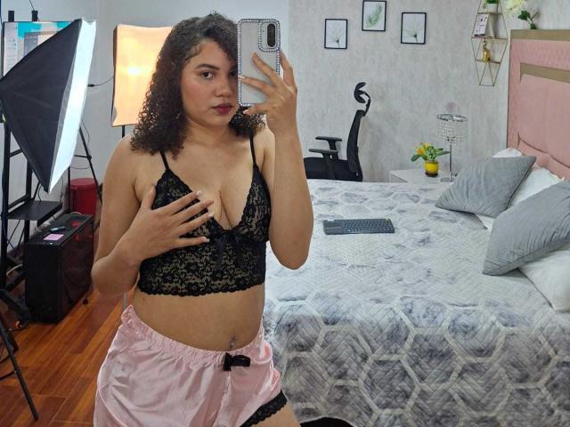 AlisonRed with curly brown hair, wearing black lace bra and pink satin shorts, covering her tits with one hand, taking a mirror selfie in a bedroom.