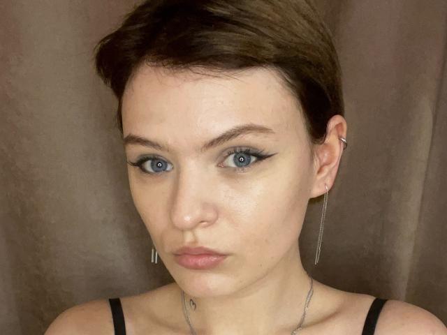 ElliLyi has short brown hair, blue eyes, and wears black eyeliner with winged tips. She has small tits, a silver necklace, and earrings. Her face is slightly flushed.