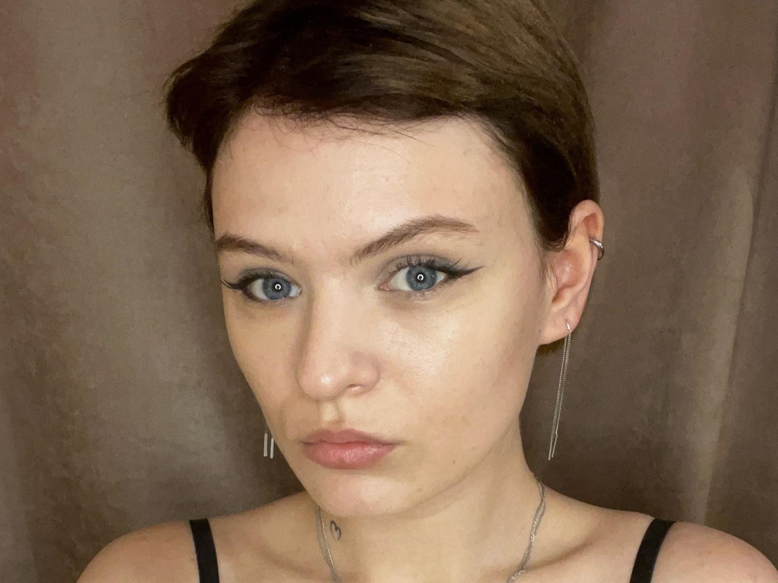 ElliLyi has short brown hair, blue eyes, and wears black eyeliner with winged tips. She has small tits, a silver necklace, and earrings. Her face is slightly flushed.