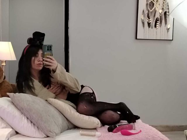 AnyuWu, com cabelo preto, usando um chapéu alto e meias pretas, está deitada em uma cama, tirando uma selfie, mostrando seus seios e vulva através de seu vestido, com um brinquedo sexual por perto..