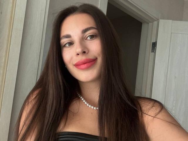 BlackFatal, com longos cabelos castanhos, batom vermelho e um colar de pérolas, sorrindo para a câmera. Ela está vestindo uma blusa preta sem alças, que destaca seus seios..