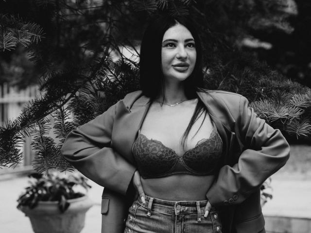 Black-and-white photo of CumAnnietime with hands on hips, wearing lace bra, denim jacket, and jeans, standing in front of pine tree, looking slightly up and to the side.