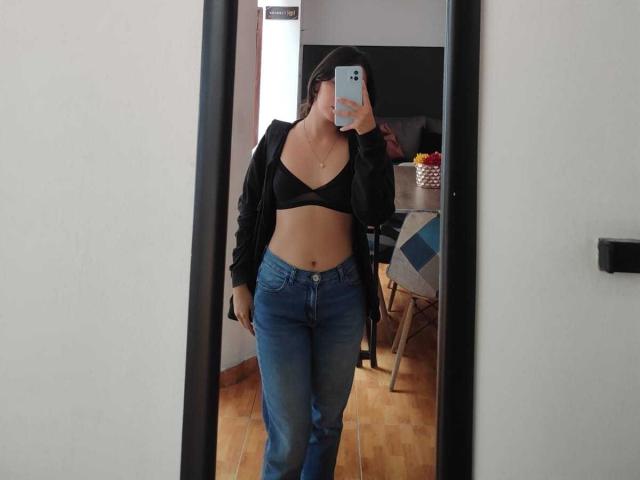 LanaKlaire è in un selfie allo specchio, indossa un reggiseno nero, una giacca nera aperta e jeans blu, mostrando il suo ventre piatto e i suoi piccoli seni, con un telefono che le copre il viso..